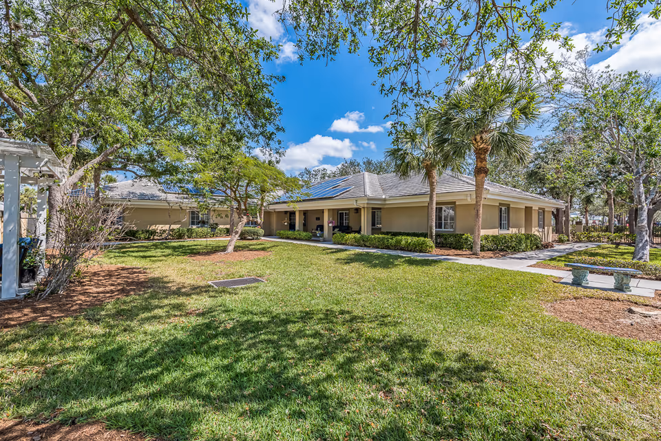 A single-story building with beige walls and a gray roof surrounded by a well-maintained grassy lawn and various trees including palm trees under a partly cloudy blue sky.