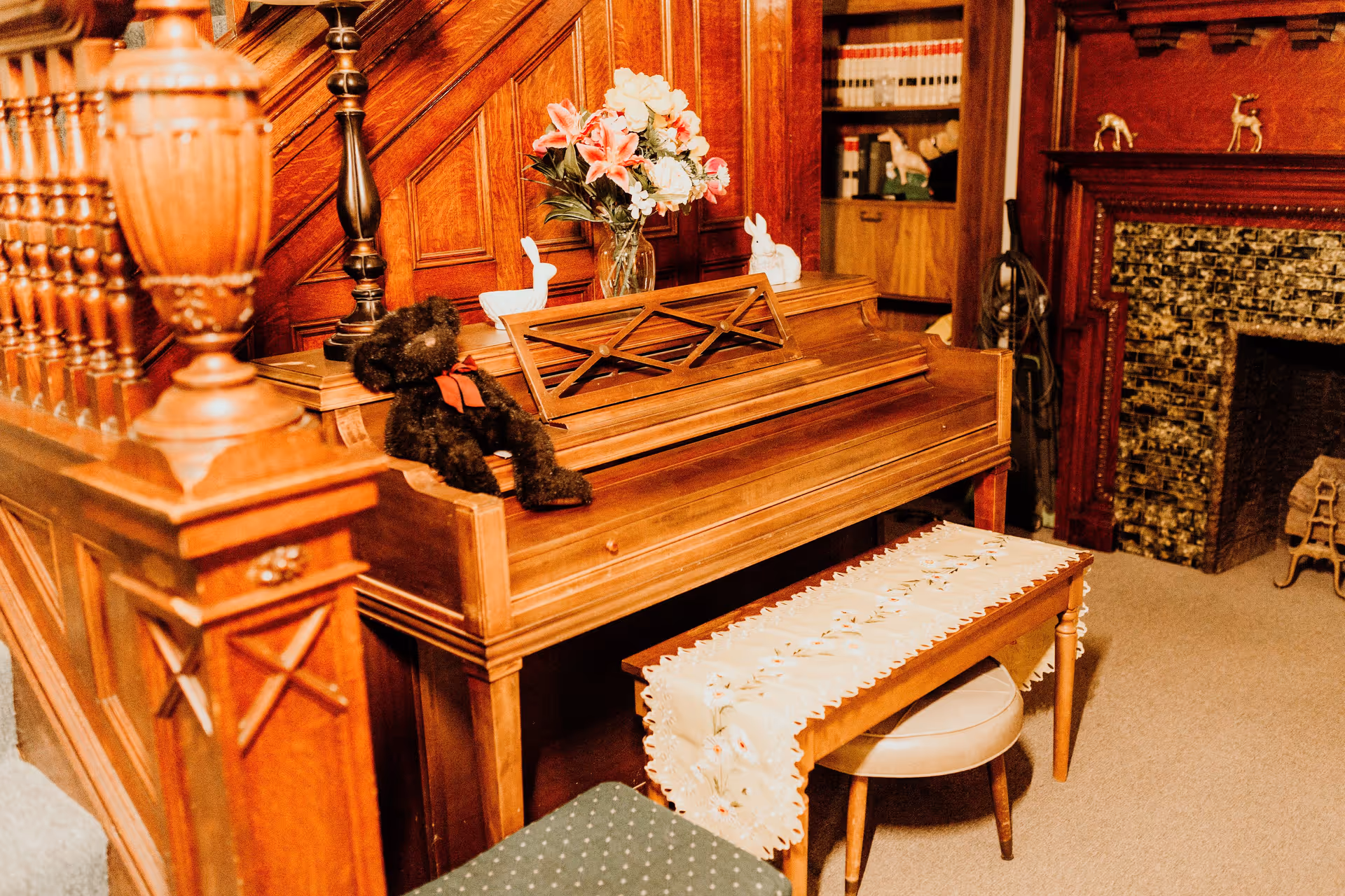 A cozy interior corner featuring a wooden piano with a floral table runner on the bench, a black teddy bear with a red bow sitting on the piano, and a vase of flowers on top. The background includes wooden paneling, a fireplace with decorative tiles, and shelves with books and small animal figurines.