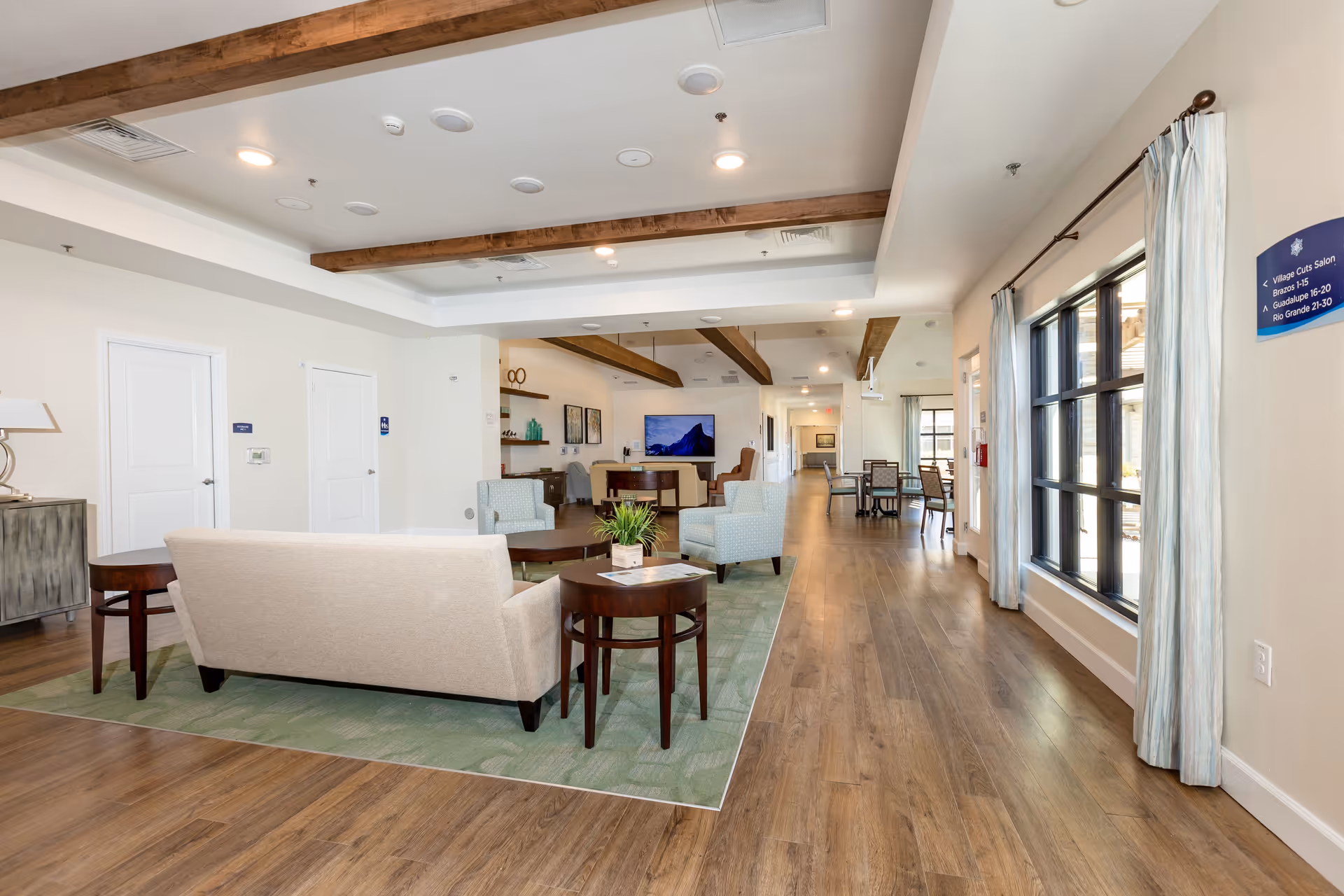 A spacious and well-lit senior living facility common area with wooden floors, beige and light blue upholstered chairs, a beige sofa, wooden side tables, and a green area rug. The ceiling features wooden beams and recessed lighting. There is a flat-screen TV mounted on the far wall, decorative shelves, and large windows with light curtains letting in natural light. A hallway extends to the right with additional seating and tables.