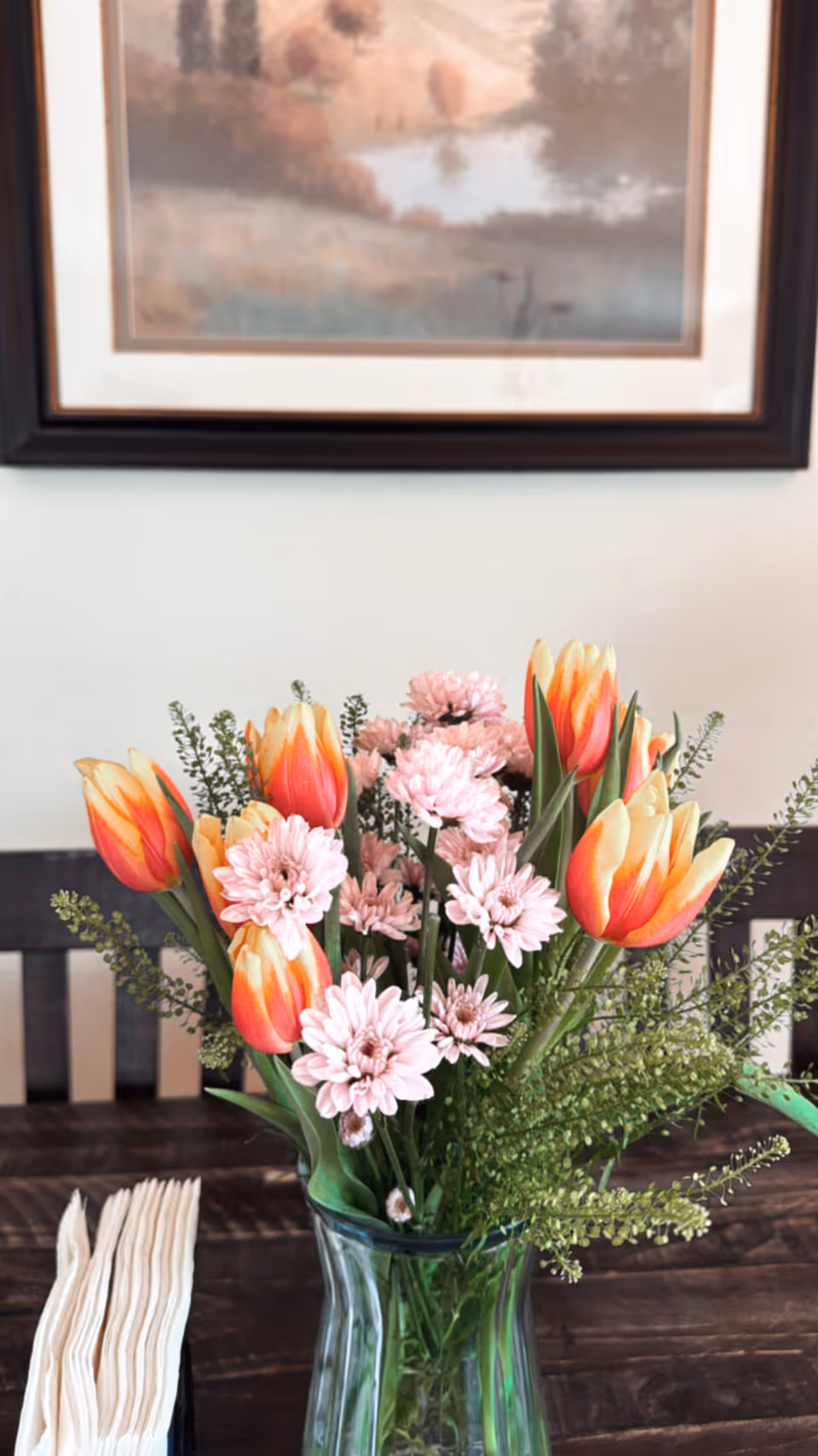A clear glass vase filled with a bouquet of orange and yellow tulips, light pink daisies, and green foliage sits on a dark wooden table. Next to the vase is a stack of white napkins. In the background, there is a framed painting of a serene landscape with trees and a body of water.
