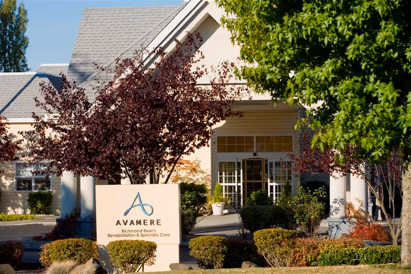 Front entrance of Avamere Richmond Beach Rehabilitation & Specialty Care with a sign, shrubs, and trees in the foreground.