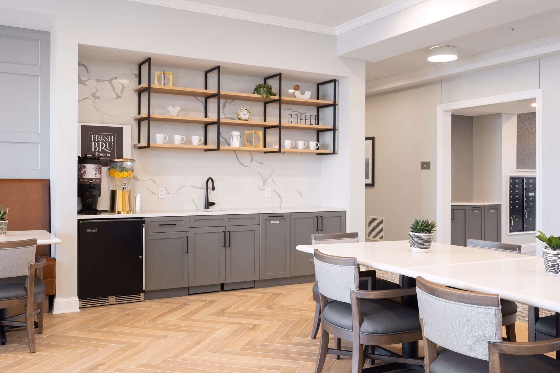 A modern dining area with a long white table surrounded by chairs. To the left, there is a countertop with gray cabinets below and open wooden shelves above holding cups, decorative items, and a sign that says 'COFFEE'. A beverage dispenser with lemon water and a coffee machine are on the counter. The floor has a light wood herringbone pattern.