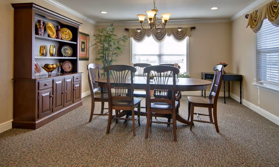 A dining room with a wooden oval table surrounded by six wooden chairs. A chandelier with multiple lights hangs above the table. There is a large window with blinds and decorative valances letting in natural light. To the left, there is a wooden cabinet displaying decorative plates and glassware. The room has beige walls and carpeted flooring.