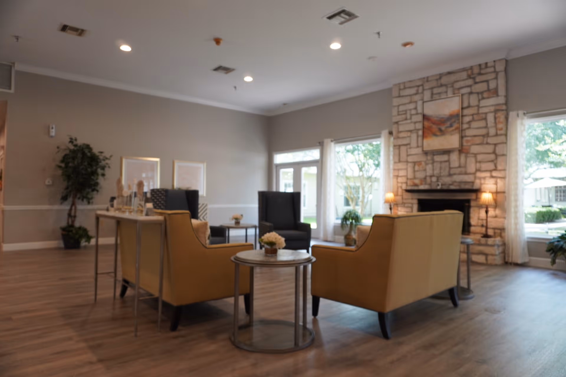 A cozy living room area with two mustard yellow armchairs and two dark gray armchairs arranged around a small round table with a flower centerpiece. The room features a stone fireplace with a painting above it, two lamps on either side of the fireplace, large windows with white curtains letting in natural light, and wooden flooring. There are also some plants and framed pictures on the walls.