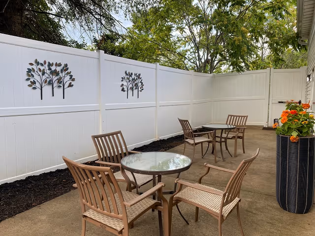 Outdoor patio area with two round glass-top tables and four brown chairs. The space is enclosed by a white fence decorated with metal tree wall art. There is a tall black planter with orange flowers on the right side and trees visible above the fence.