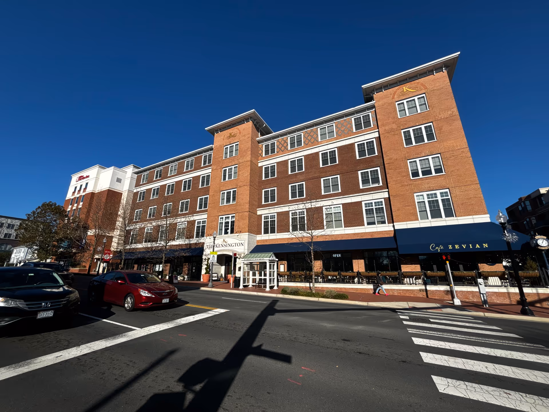Front view of The Kensington Falls Church, a multi-story brick senior living building with storefront awnings at a street intersection under a clear blue sky.