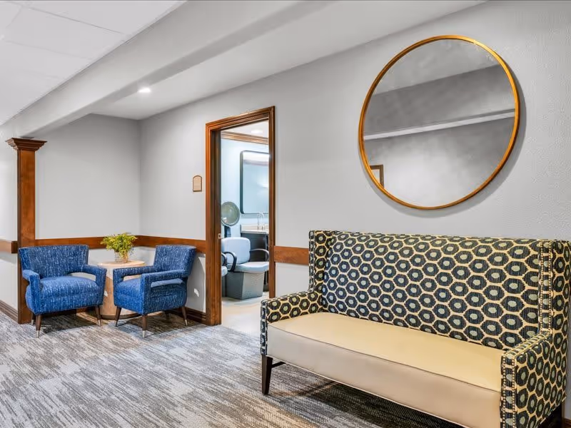 A cozy seating area in a senior living facility with two blue upholstered armchairs and a small round table with a plant between them, a patterned loveseat, and a large round mirror on the wall. A doorway leads to another room with additional seating and a sink.