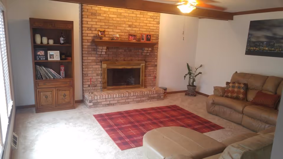 A cozy living room with a brick fireplace in the center, a wooden shelf with decorative items and books to the left, a red plaid rug on the carpeted floor, a beige leather sofa with patterned cushions on the right, a potted plant in the corner, and a ceiling fan with a light above.