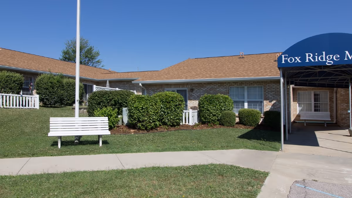 Front exterior of a single-story brick senior living building with shrubs, white benches, a flagpole, and a blue entrance awning.