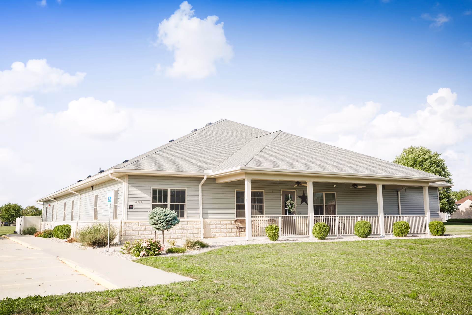 Single-story building with light gray siding and a covered porch with railings. The building is surrounded by a well-maintained lawn and some small shrubs. The sky is clear with a few clouds.