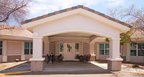 Entrance of a single-story building with a covered driveway supported by columns, beige walls, and a tiled roof. There are potted plants and chairs near the double glass doors under the covered area. Trees with sparse leaves are visible on either side of the building.
