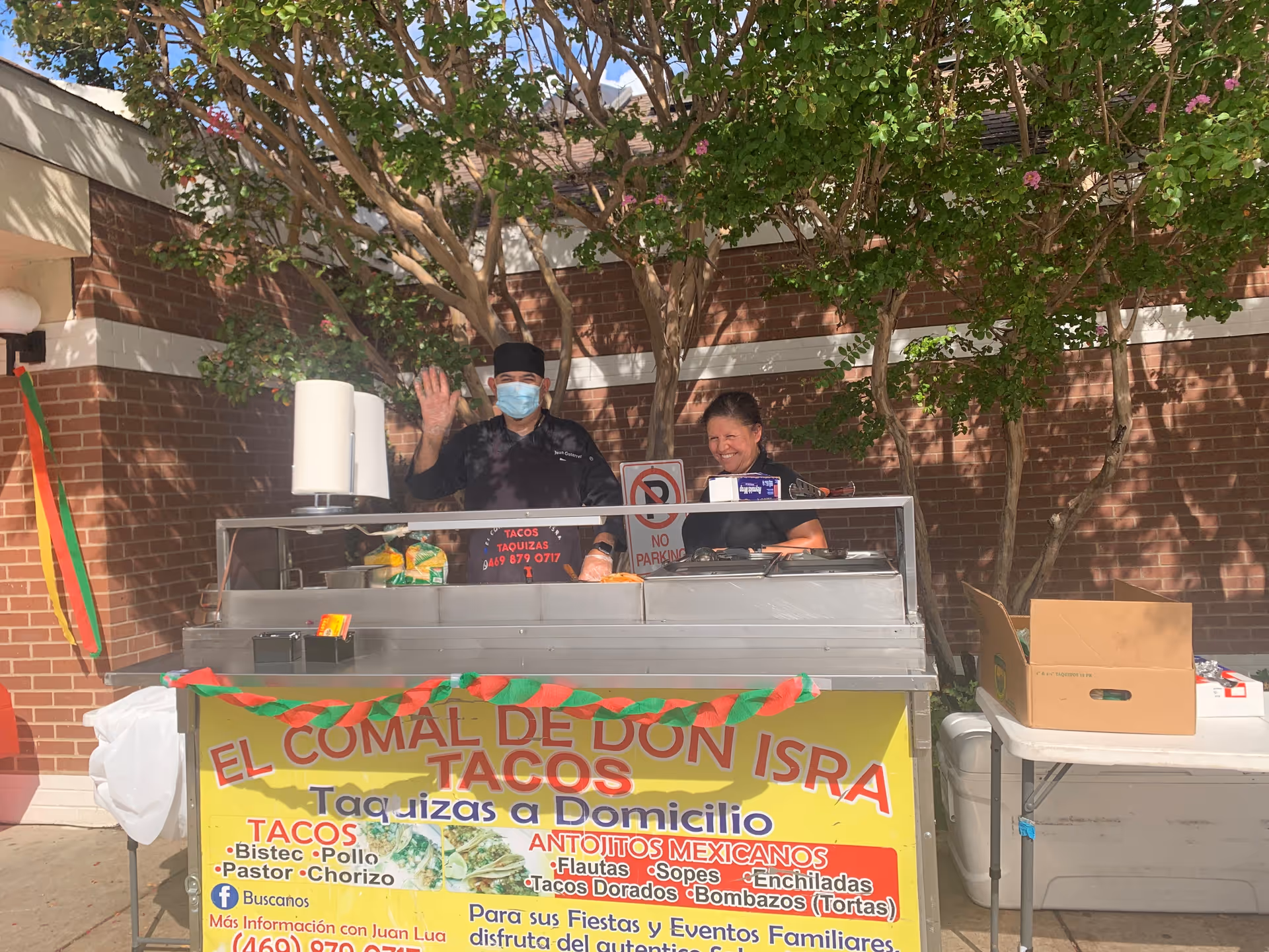Two workers stand behind a decorated taco cart outdoors in front of a brick wall and trees, one waving and wearing a face mask.