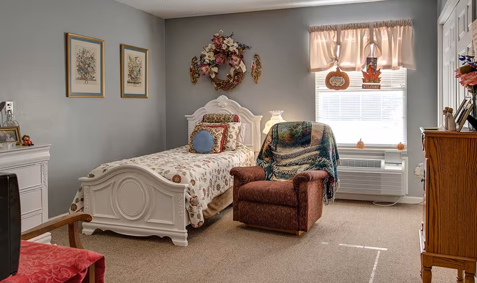 A cozy bedroom with a single white ornate bed covered with a floral quilt and decorative pillows. Next to the bed is a brown upholstered armchair draped with a colorful blanket. The room has light gray walls adorned with two framed floral prints and a decorative wreath above the bed. A window with a pink valance lets in natural light, and a wooden dresser with flowers and picture frames is visible on the right side.