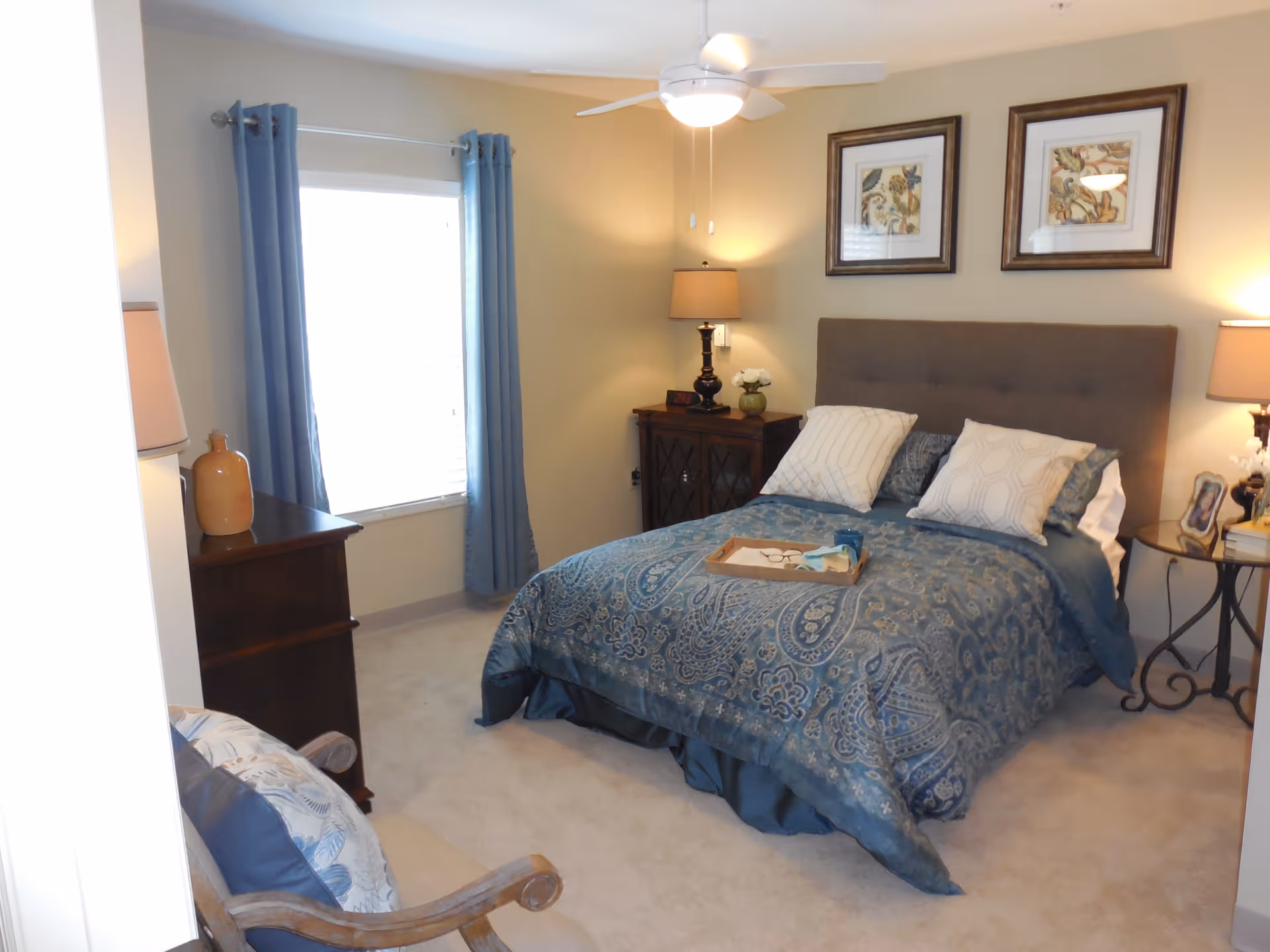 A cozy bedroom with a neatly made bed featuring a blue patterned comforter and multiple pillows. There are two framed artworks hanging above the bed. On either side of the bed are nightstands with lamps, a clock, and decorative items. A window with blue curtains lets in natural light, and a wooden chair with cushions is positioned near the entrance.