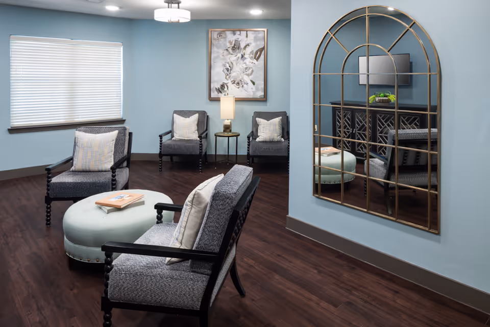 A cozy sitting area with four cushioned chairs arranged around a round, light green ottoman with a book on top. The room has light blue walls, dark wood flooring, a large decorative mirror on one wall, a window with closed blinds, a small side table with a lamp, and a floral painting hanging above two chairs.