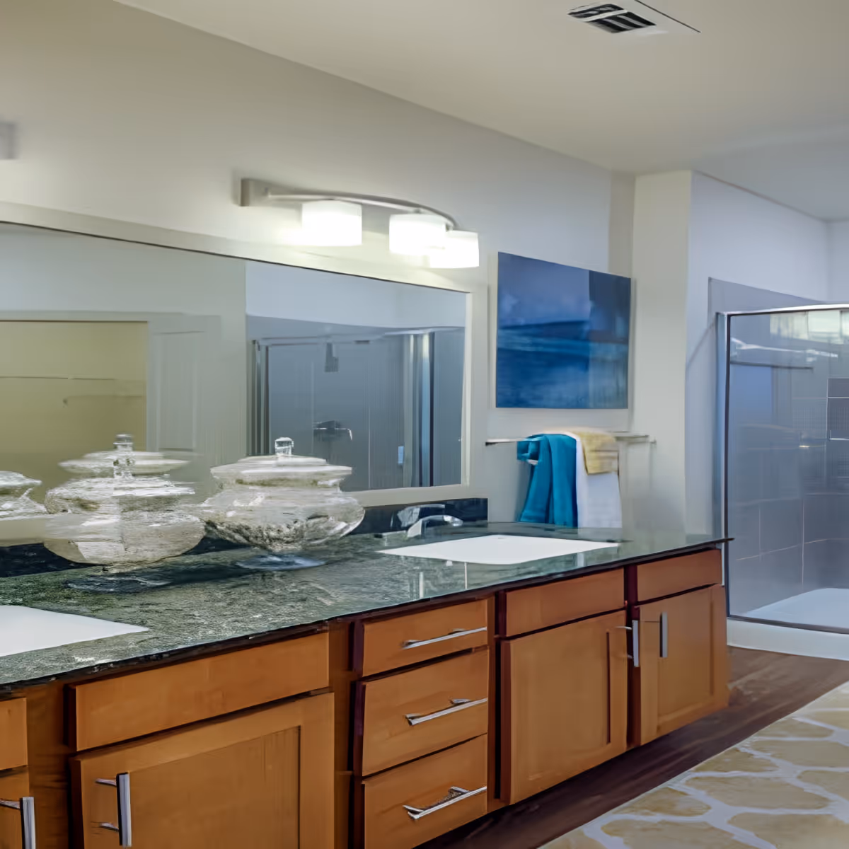 Modern bathroom with a long granite countertop featuring two sinks and wooden cabinets underneath. Above the countertop is a large mirror with three light fixtures. On the right side, there is a glass-enclosed shower and a towel rack with blue and white towels. A blue abstract painting hangs on the wall above the towel rack.