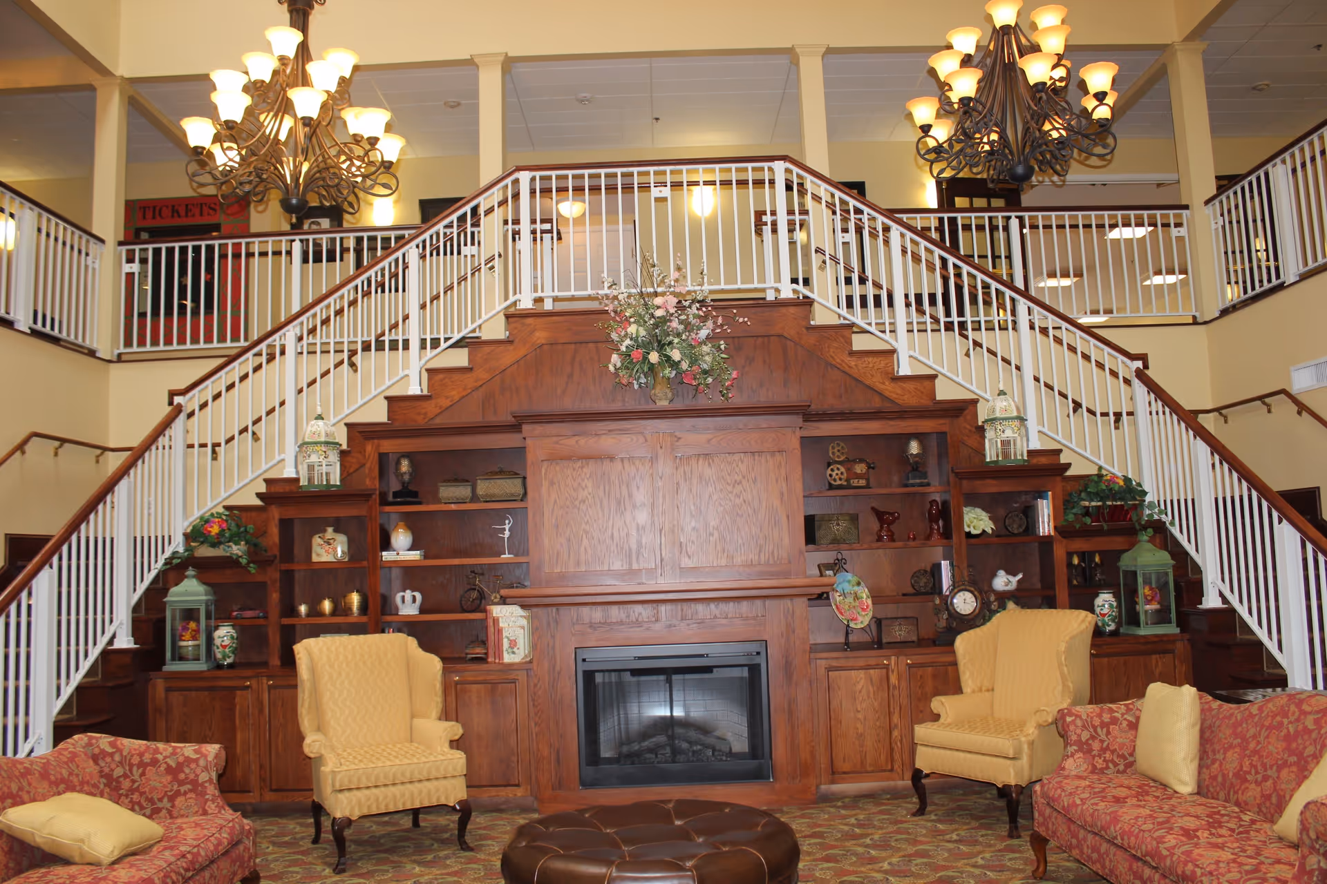A spacious senior living community lounge area featuring a large wooden built-in bookshelf and fireplace under a staircase with white railings. The room is furnished with yellow armchairs, red patterned sofas with yellow cushions, a round brown ottoman, and decorated with floral arrangements and various decorative items. Two large chandeliers hang from the ceiling.