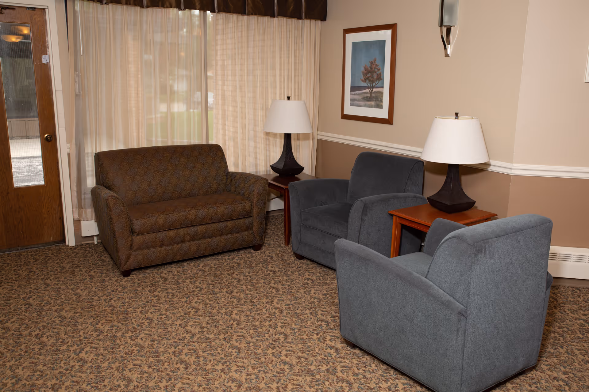 A cozy living room area with a brown patterned loveseat, two gray armchairs, two wooden side tables each with a lamp, a framed picture on the wall, and a window with sheer curtains letting in natural light.