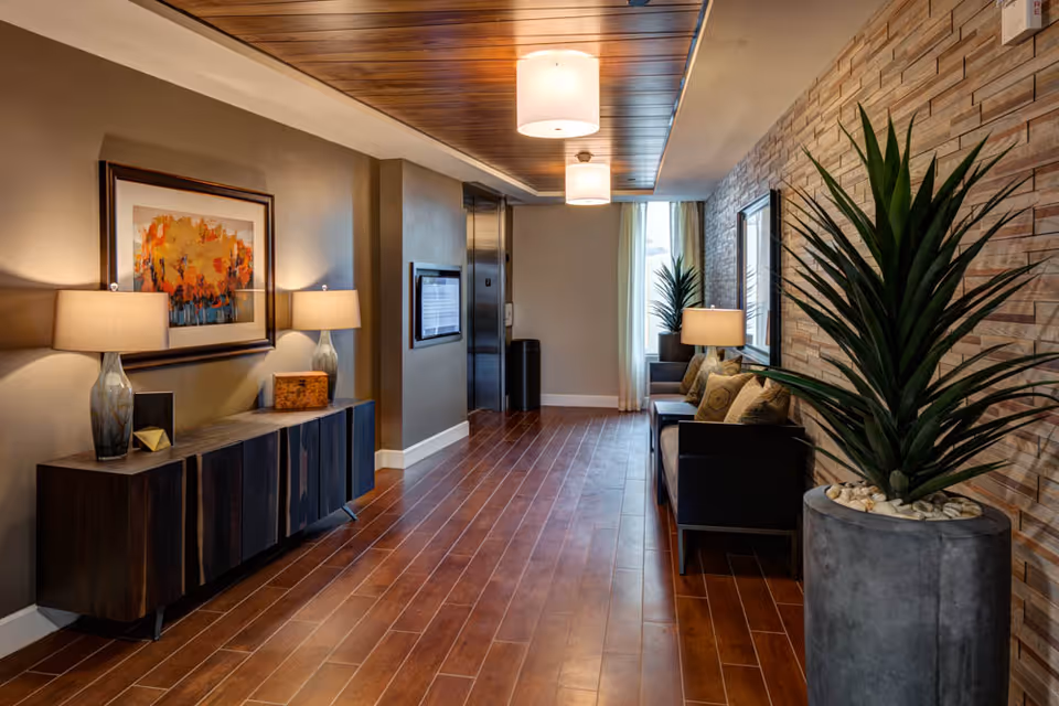 A well-lit hallway with wooden flooring and a wooden ceiling panel. On the left side, there is a dark wooden cabinet with two table lamps and a framed abstract painting above it. On the right side, there is a stone-textured wall with a large potted plant in the foreground and a seating area with a sofa, cushions, and a table lamp near a window with sheer curtains. An elevator door is visible at the end of the hallway.
