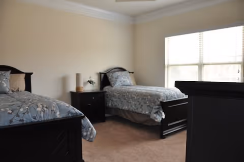 Two twin beds with matching blue floral bedding in a bright bedroom with beige walls and a large window.