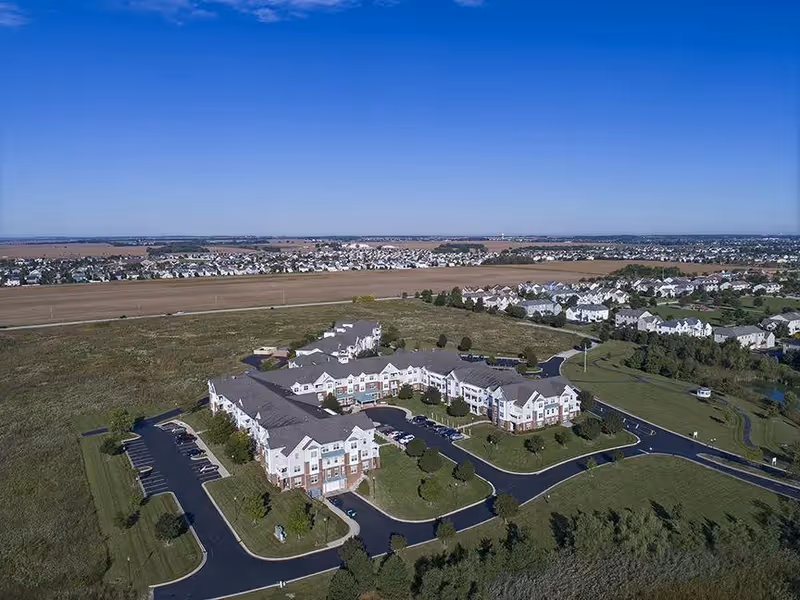 Aerial view of a large senior living facility named American House Cedarlake surrounded by green lawns and parking lots, with a suburban neighborhood and open fields in the background under a clear blue sky.