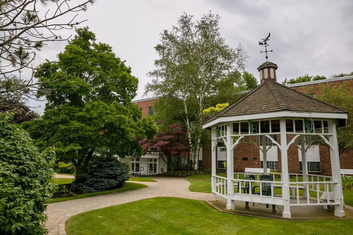 A peaceful outdoor garden area at Brentwood Rehabilitation And Healthcare Center featuring a white wooden gazebo with a weather vane on top, surrounded by green grass, trees, and a paved walkway leading to a brick building in the background.