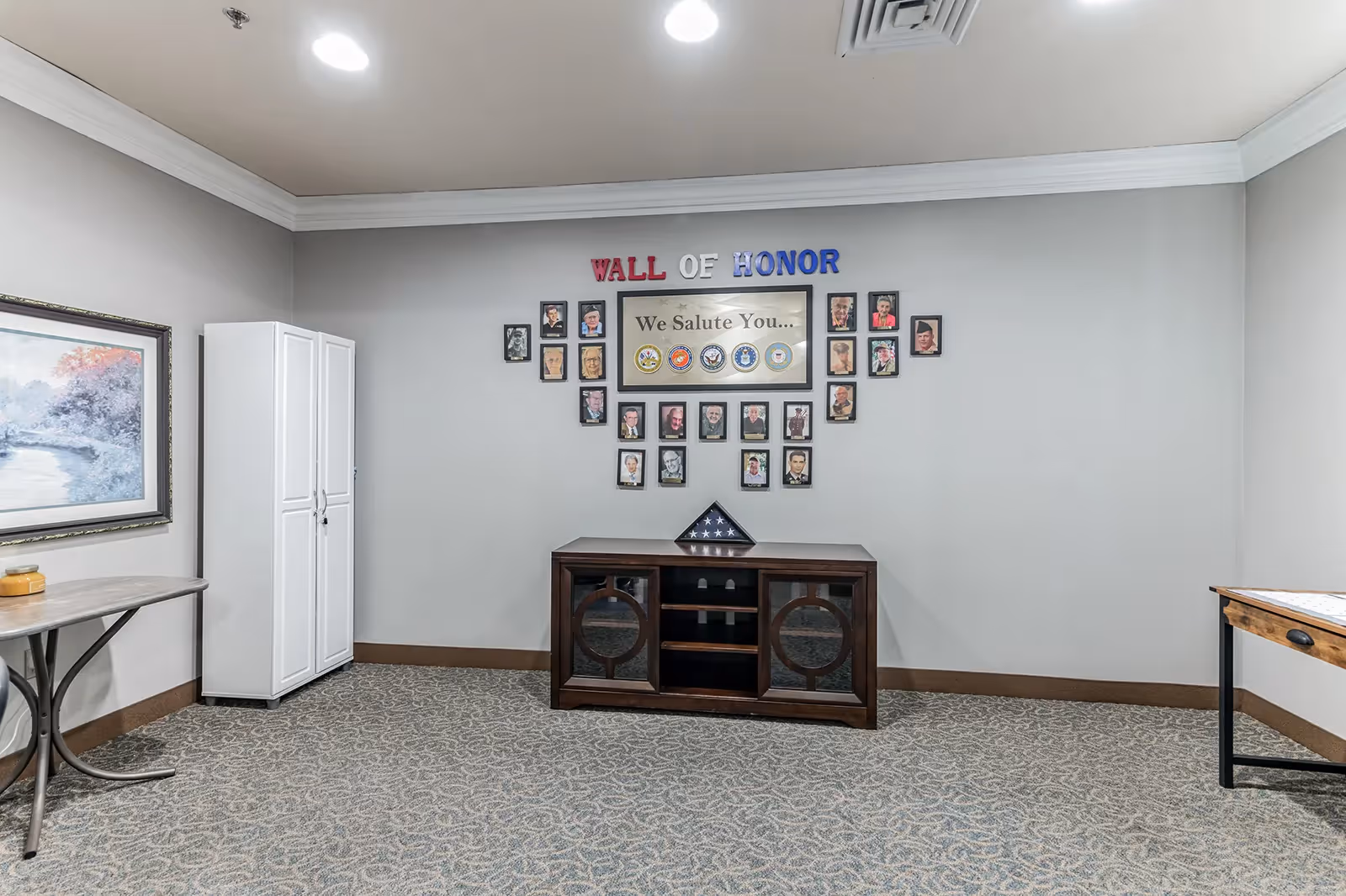Interior room showing a 'Wall of Honor' photo display above a wooden cabinet with framed artwork and tables on either side.