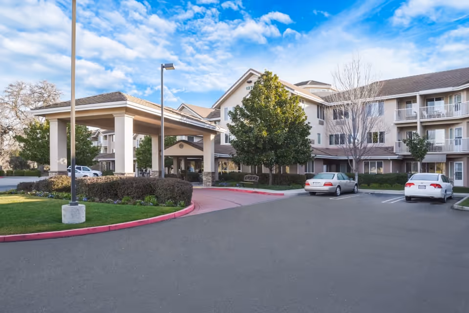 Multi-story senior living building front with a covered porte-cochere, landscaped entrance and parked cars.