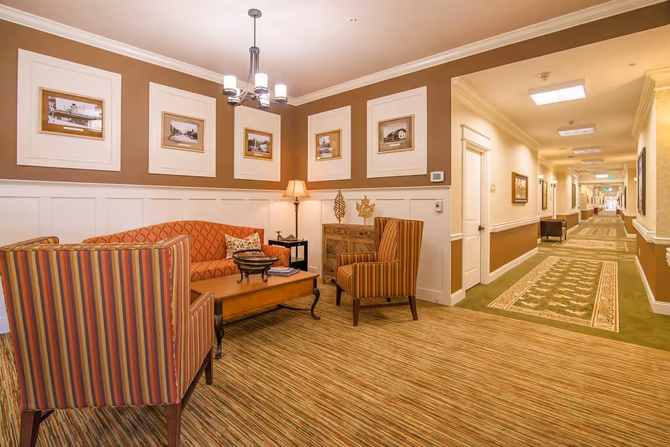 A cozy seating area in a senior living facility hallway with a striped armchair, an orange patterned sofa, a wooden coffee table, a floor lamp, and framed black and white photos on the walls. The hallway extends to the right with carpeted floors and additional seating visible in the distance.