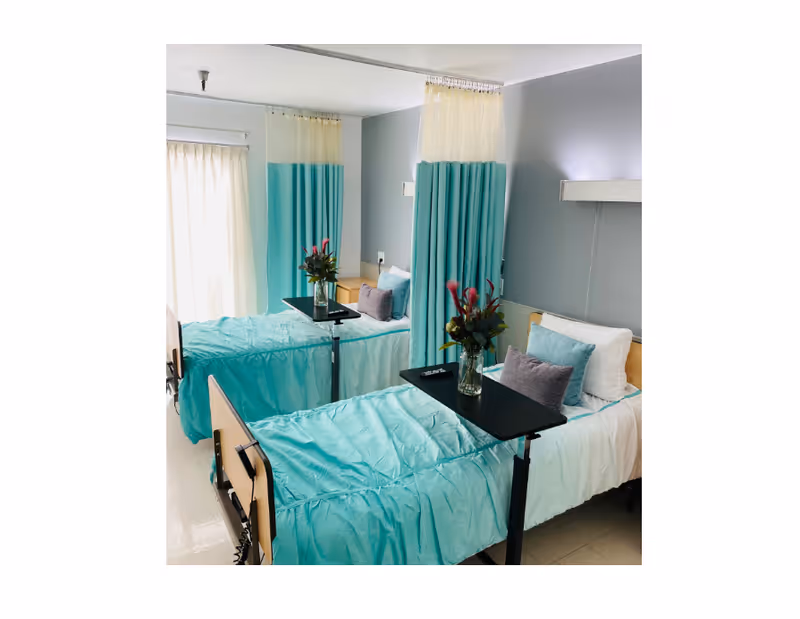 Two hospital-style beds with turquoise bedding and pillows separated by a turquoise privacy curtain in a well-lit room. Each bed has a black overbed table with a vase of flowers on it. The room has light-colored walls and a window with sheer curtains.