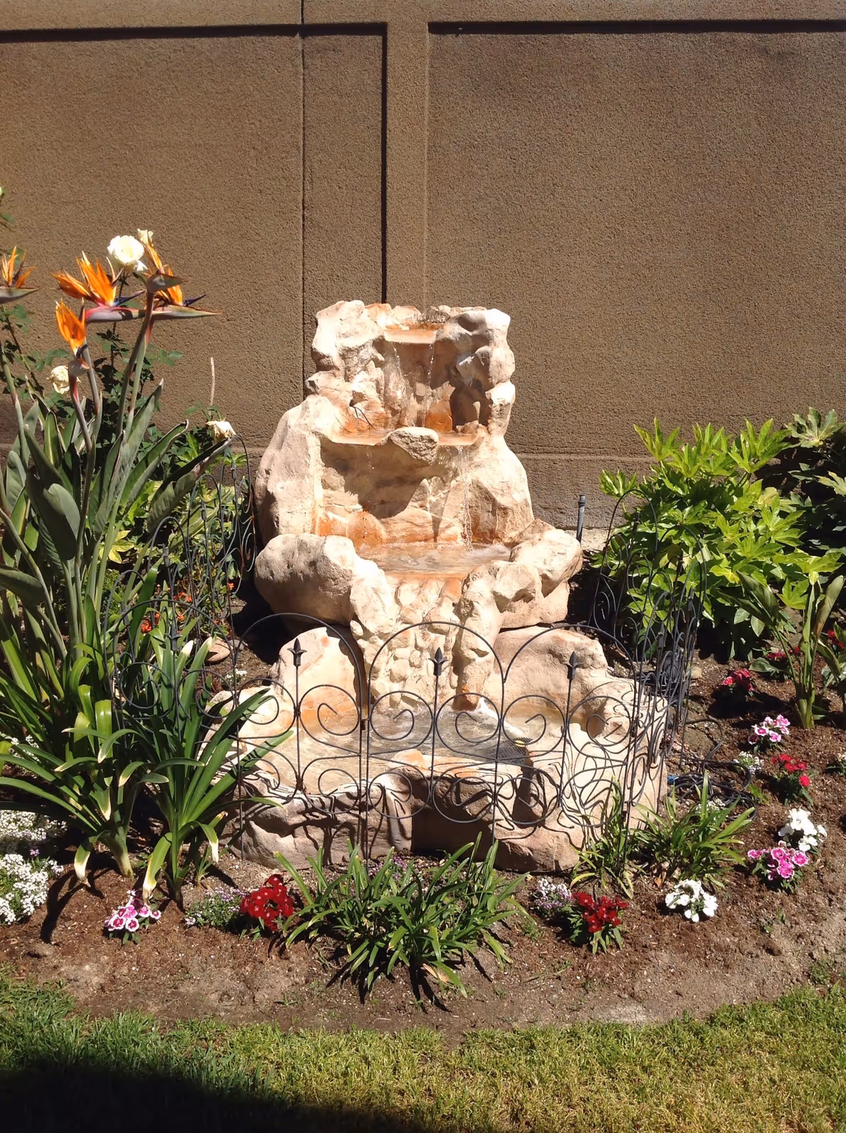 A small outdoor garden area featuring a stone water fountain surrounded by various green plants and colorful flowers, with a beige wall in the background.