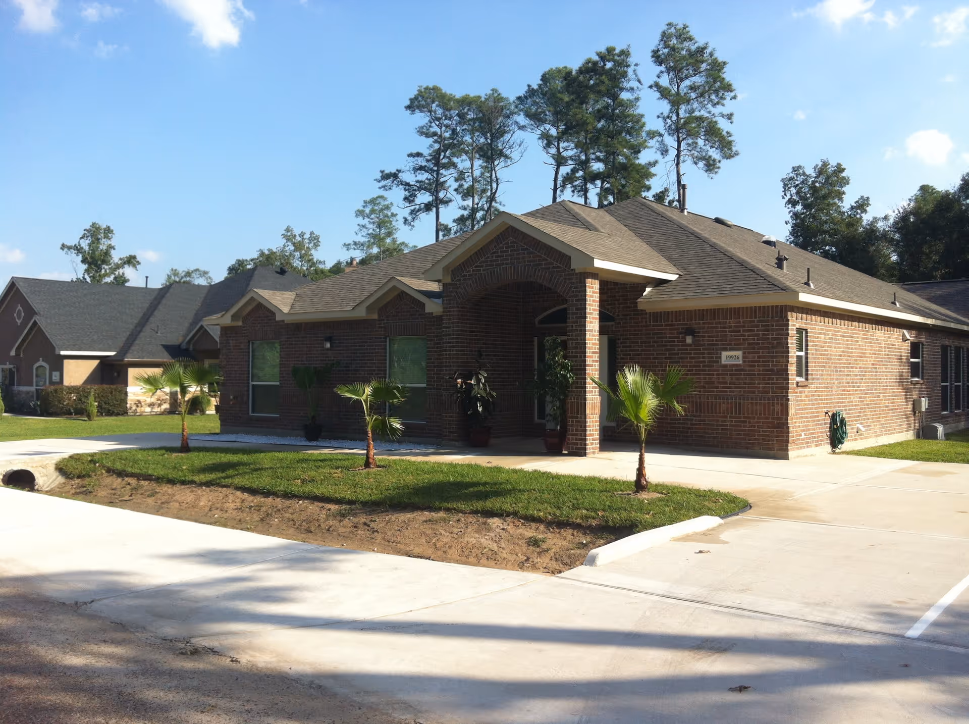 Exterior view of a single-story brick building with a covered entrance, small palm trees planted in front, and a concrete driveway. Tall trees and a clear blue sky are visible in the background.