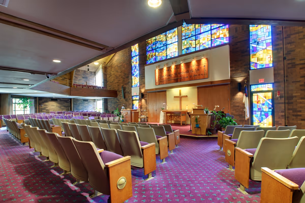 Interior view of a chapel with rows of beige and purple cushioned chairs facing a wooden altar with a cross. The chapel features stained glass windows with colorful geometric patterns and brick walls. There are plants near the altar and a purple carpet with a small pattern covering the floor.