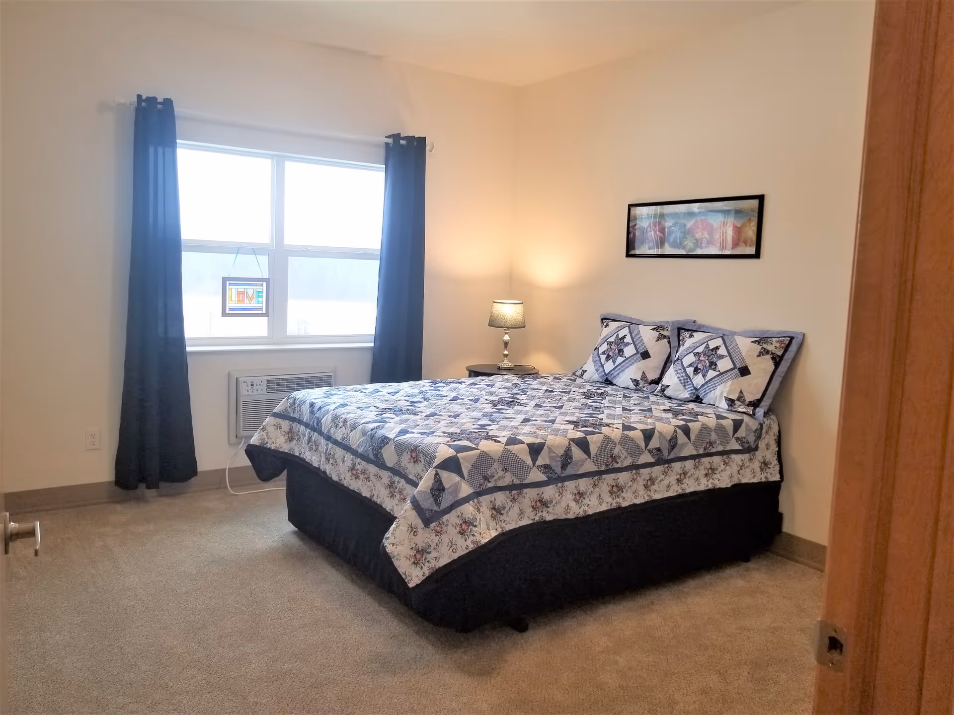 A bedroom with a bed covered in a blue and white patterned quilt and matching pillows. There is a window with dark blue curtains, an air conditioning unit below the window, a small round bedside table with a lamp, and a framed picture on the wall above the bed.