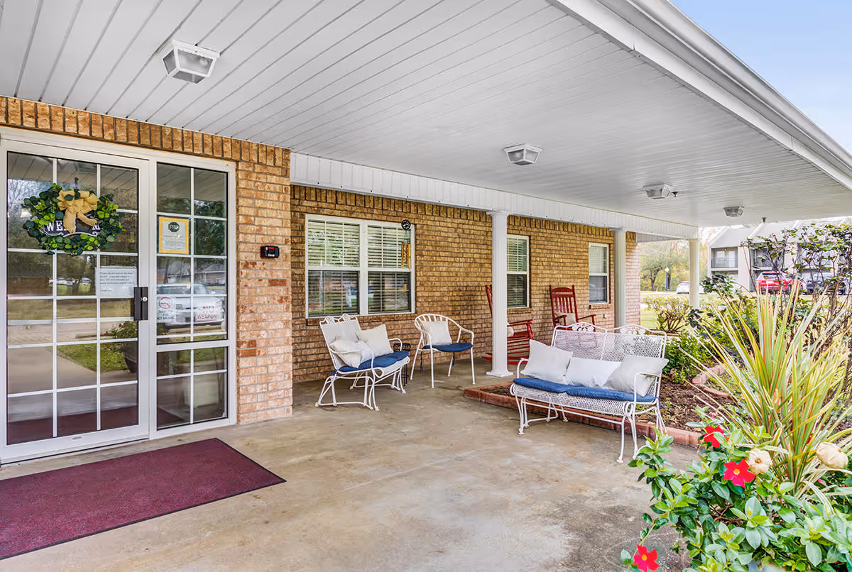 Covered outdoor patio area at Meadowview Place with brick walls, white ceiling, and several white metal chairs and benches with cushions. There is a glass door with a green wreath and a welcome sign, a maroon floor mat, and some flowering plants in the foreground.