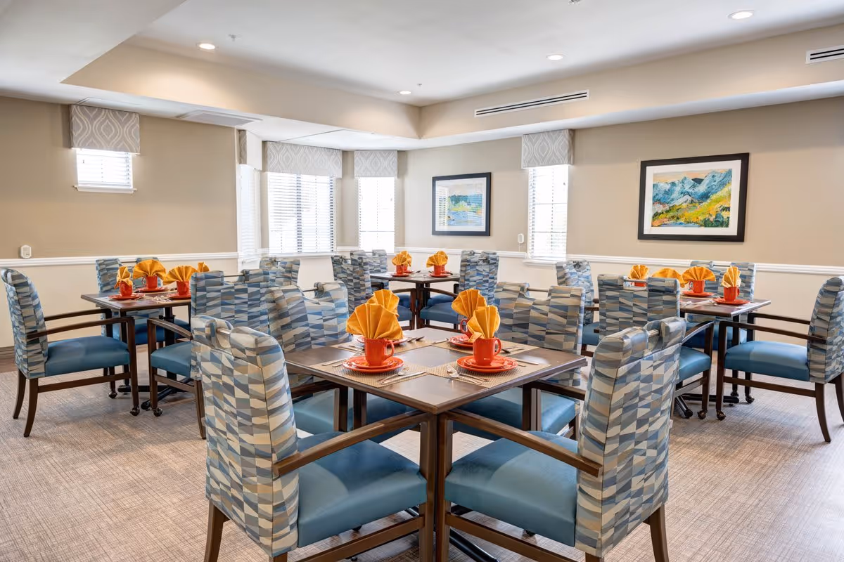 A bright dining room with multiple square tables, each set with orange cups, plates, and yellow folded napkins. The chairs have blue patterned upholstery and the room features beige walls with white trim, several windows with blinds, and framed landscape paintings on the walls.