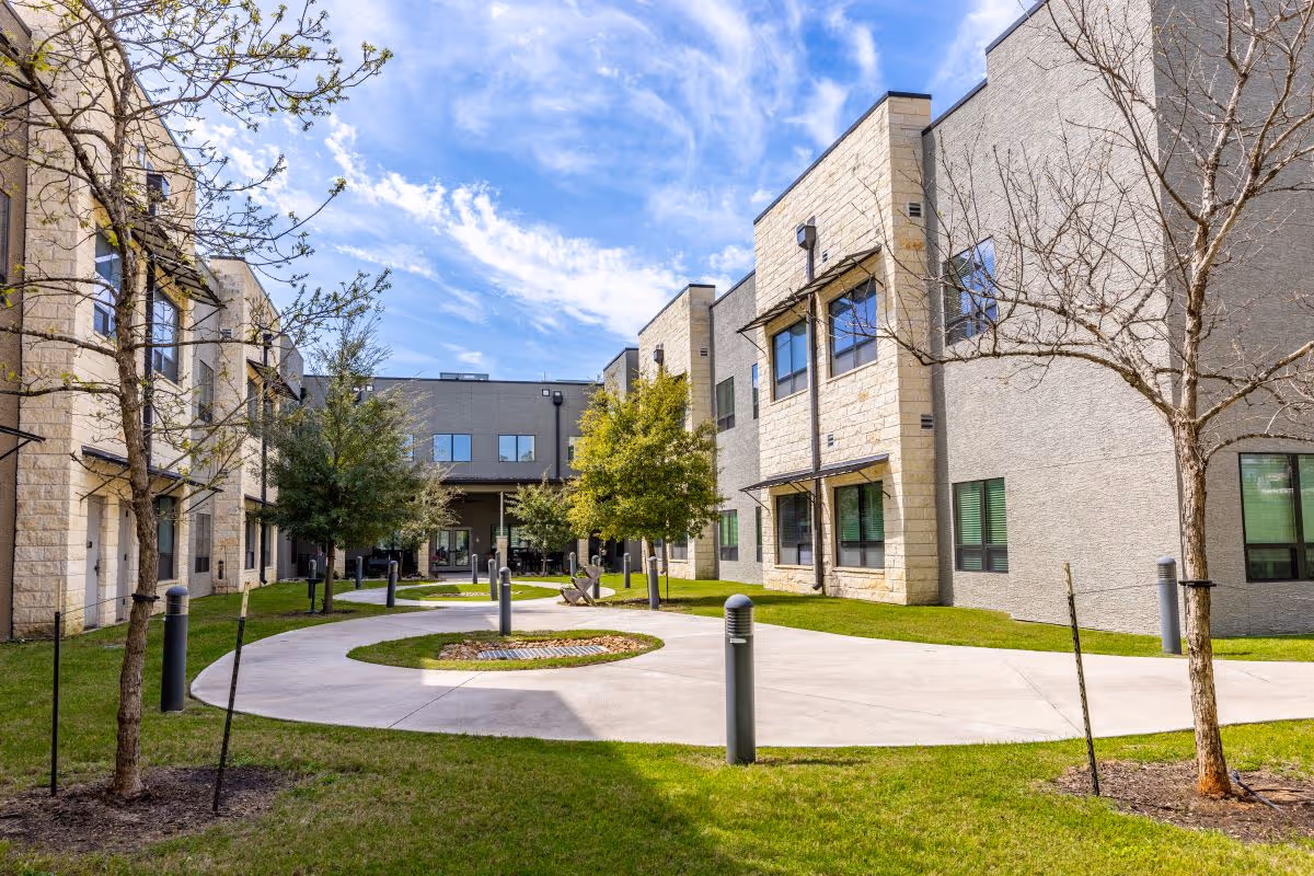 Courtyard between modern beige-stone senior living buildings with a circular concrete walkway, trees, and green lawns under a blue sky.