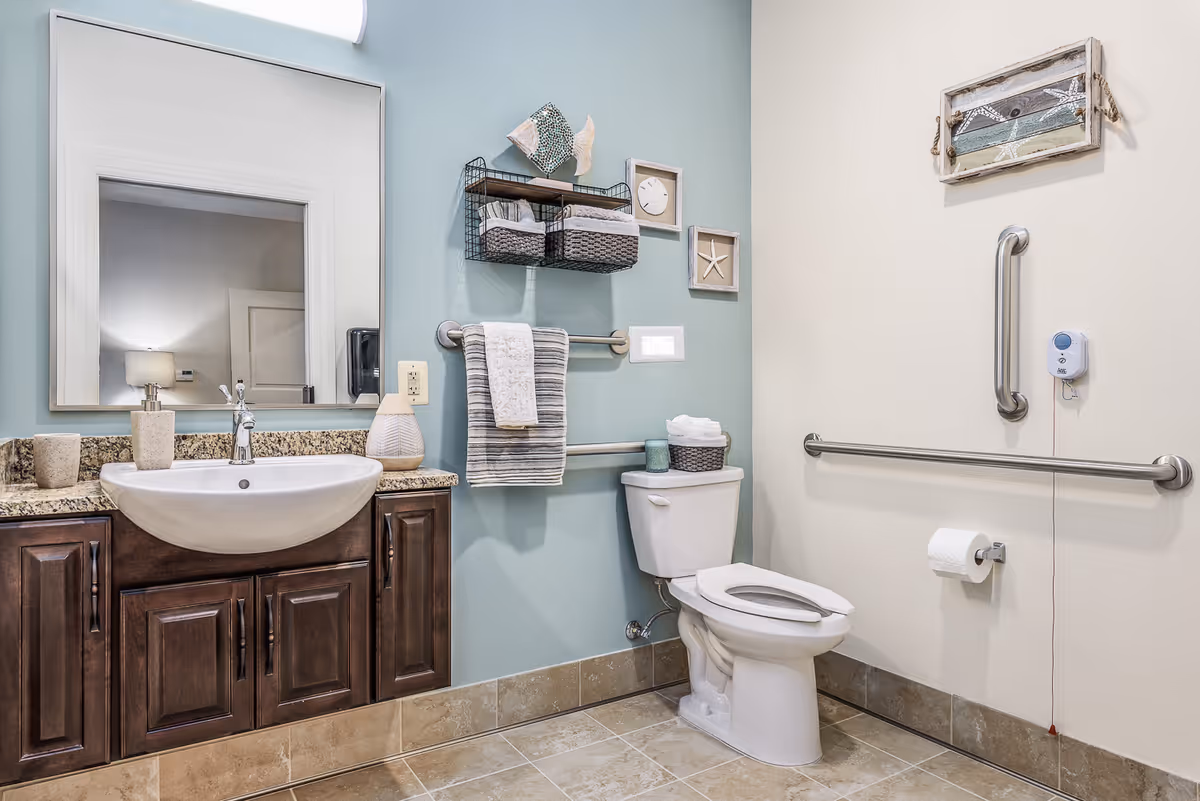 A clean and accessible bathroom with a white toilet and a sink with a granite countertop and dark wooden cabinets. The walls are painted in light blue and beige, decorated with small framed art pieces and a wire basket holding towels. There are metal grab bars installed near the toilet for support, and a call button is mounted on the wall. The floor is tiled in a beige color.