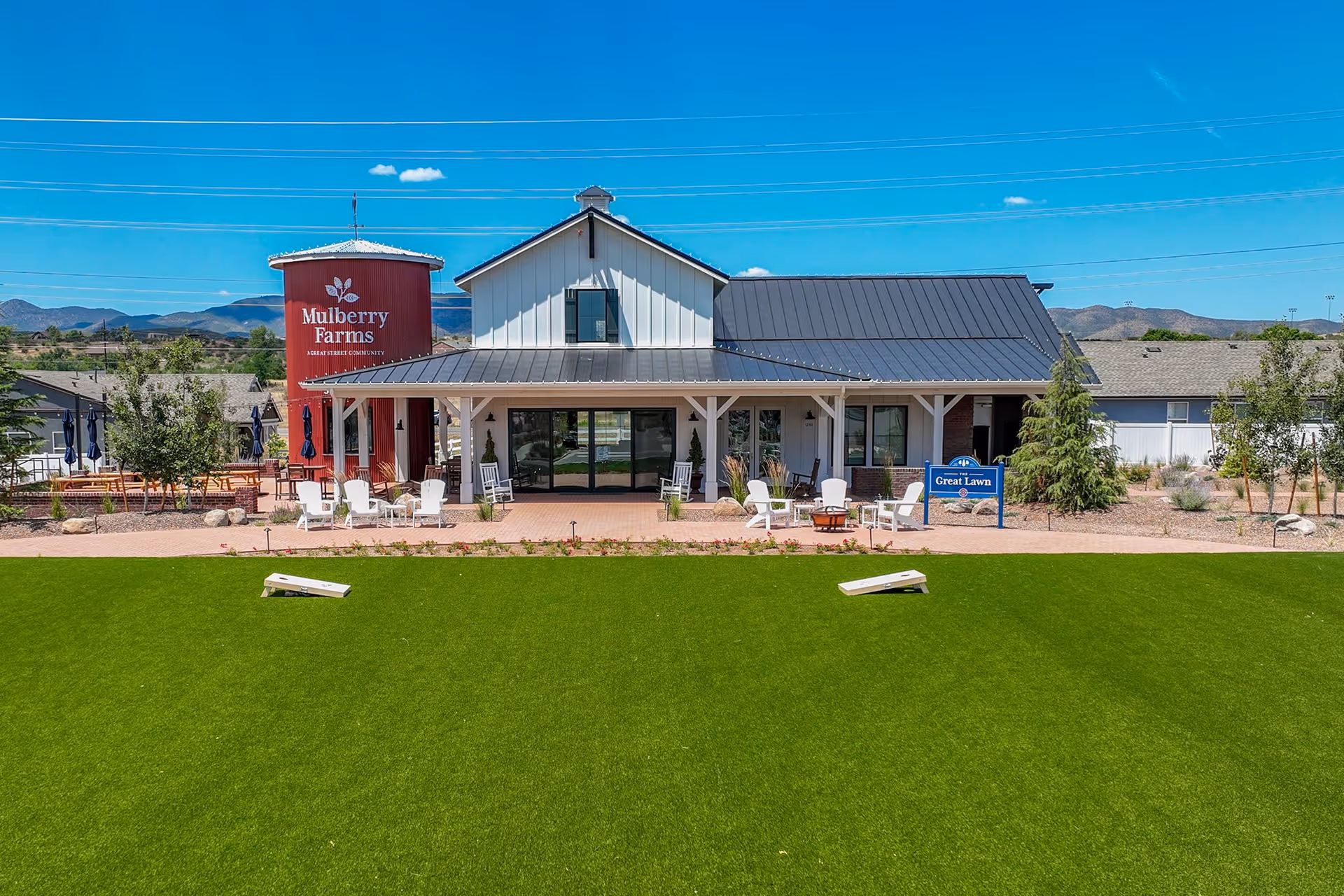 Outdoor view of Mulberry Farms facility showing a white building with a black roof and a red silo with the Mulberry Farms logo. In front of the building is a large green lawn with two cornhole boards, several white chairs, and a fire pit. There is a sign that reads 'Great Lawn' near the seating area. The background includes mountains and a clear blue sky.