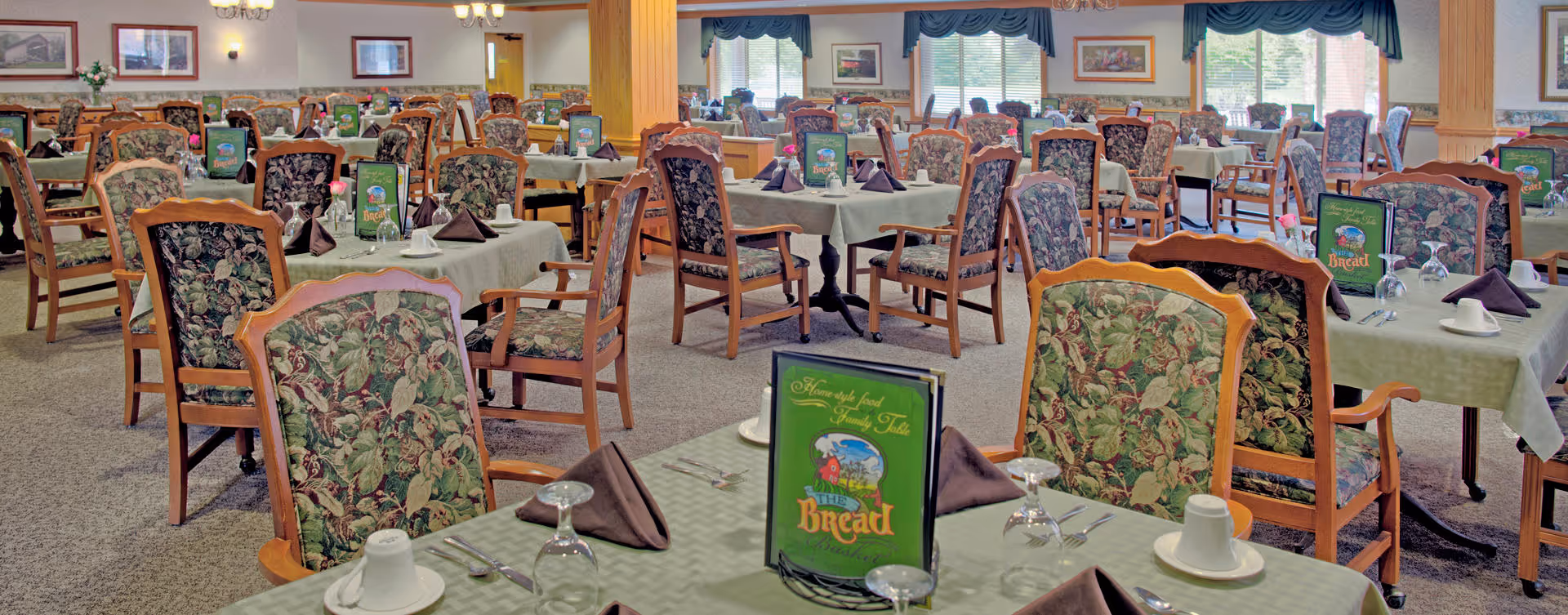 A spacious dining room with multiple tables covered in light green tablecloths, each set with brown folded napkins, white cups, glasses, and silverware. The chairs have wooden frames with floral-patterned upholstery. The room is well-lit with large windows covered by blue valances and framed artwork on the walls.