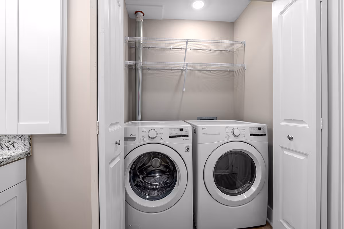 A laundry area with a front-loading washing machine and dryer side by side, enclosed by white bi-fold doors. Above the machines are two white wire shelves mounted on the beige wall. To the left, part of a white cabinet and granite countertop is visible.
