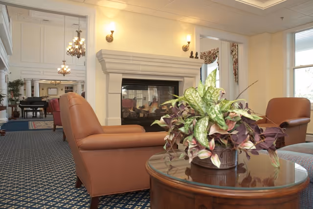 A cozy living room area with two brown leather armchairs facing a white fireplace. A round wooden table with a potted plant is in the foreground. In the background, there is a grand piano and more seating, with chandeliers hanging from the ceiling and large windows letting in natural light.