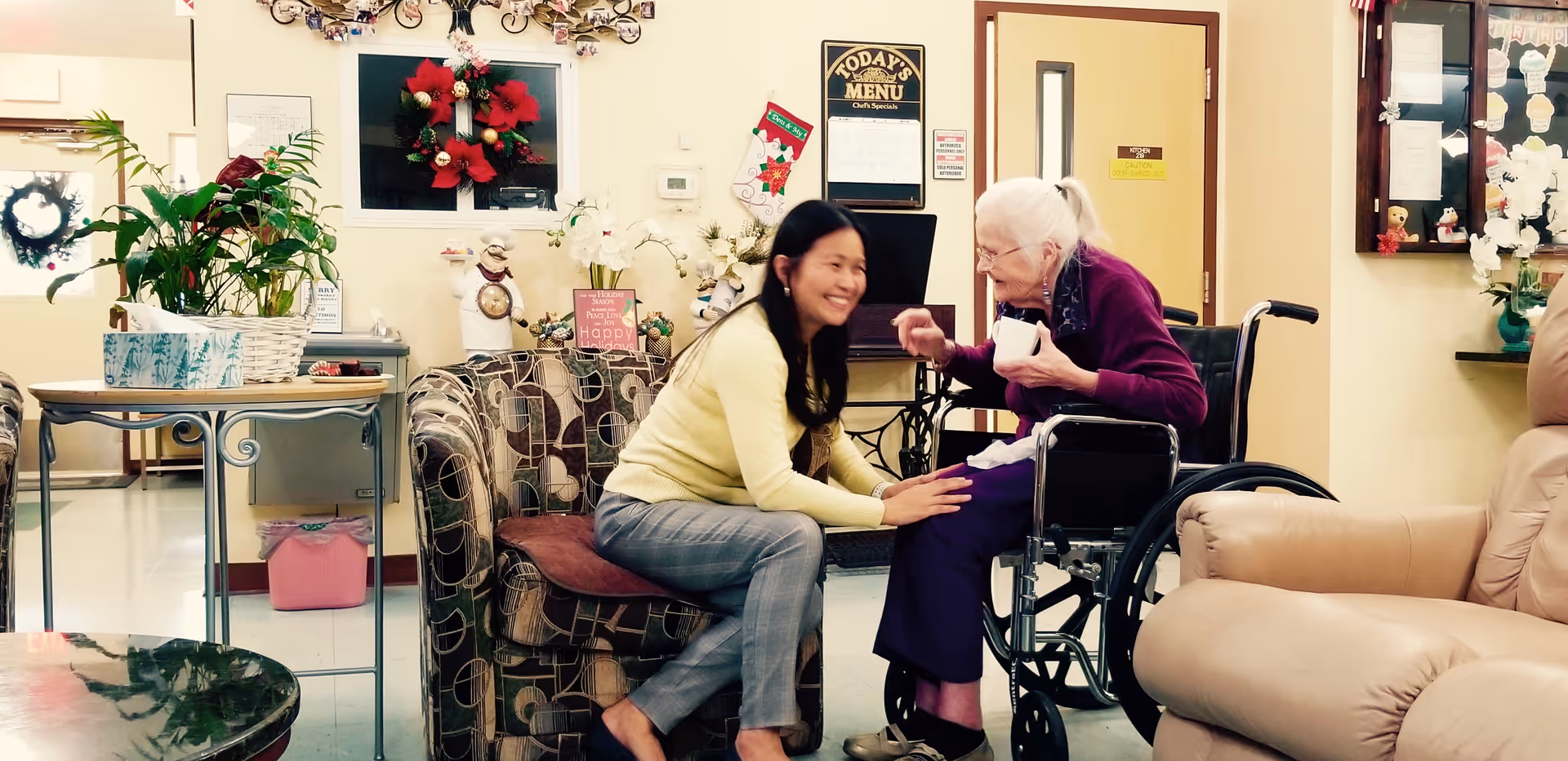 An elderly woman in a wheelchair smiling and interacting with a younger woman who is kneeling beside her in a cozy, decorated common area of an assisted living facility. The room has comfortable chairs, plants, holiday decorations, and a menu board on the wall.