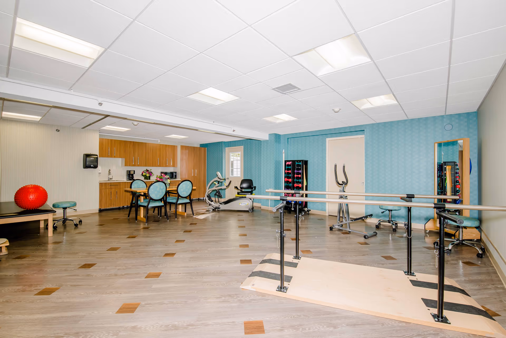 A spacious rehabilitation or physical therapy room with wooden flooring and a blue accent wall. The room contains parallel bars for walking exercises, exercise equipment including a stationary bike, a rack of dumbbells, a large mirror, a table with four chairs, and wooden cabinets with a countertop and sink. There is also a red exercise ball and a stool on the left side.