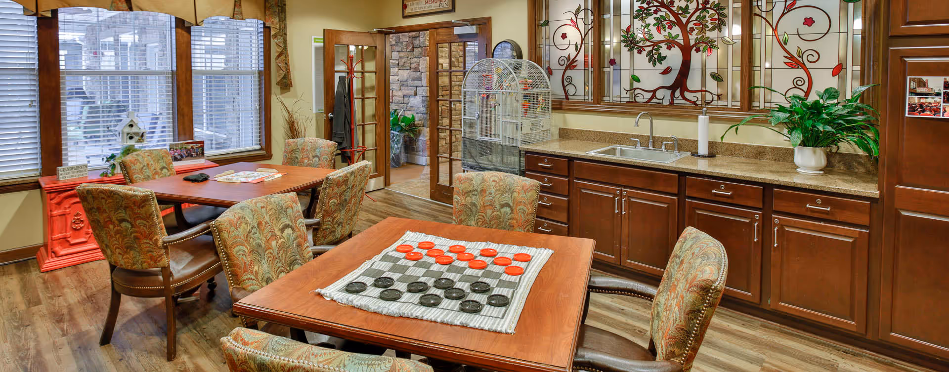 A cozy common area with wooden tables and patterned upholstered chairs. One table has a checkers game set up with red and black pieces. The room features large windows with blinds, a countertop with a sink, cabinets, and decorative wall art with floral designs. There is a birdcage on the counter and a red pedestal near the window. French doors lead to another room with stone walls and plants.