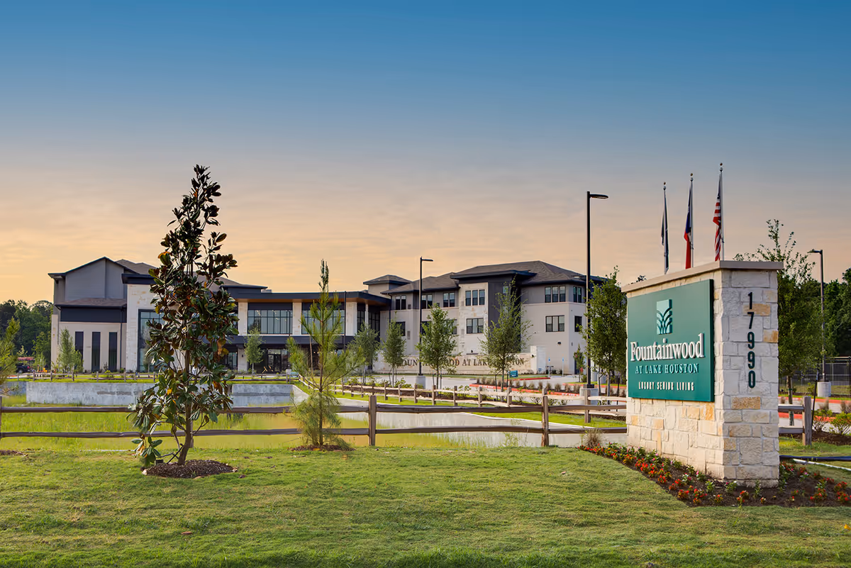 Exterior view of Fountainwood at Lake Houston senior living facility during sunset, showing a modern multi-story building with landscaped grounds, trees, and a stone sign with the facility name and address 17990.
