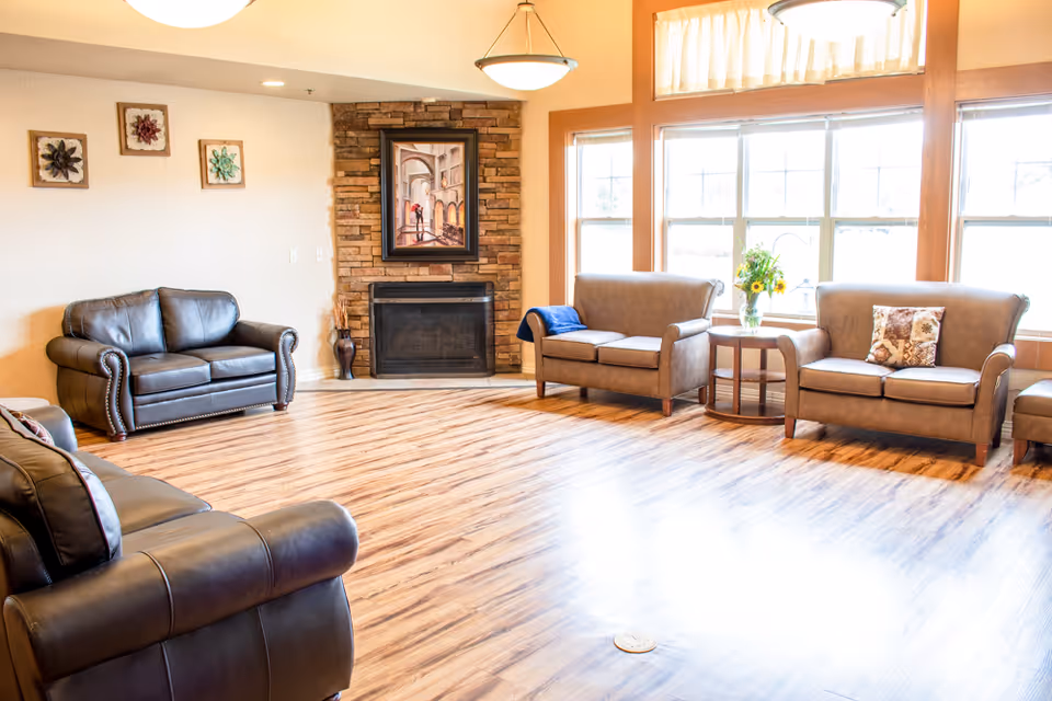 A bright and spacious living room with wooden flooring, several leather sofas arranged around a stone fireplace with a framed painting above it, large windows letting in natural light, and a small round table with a vase of flowers.