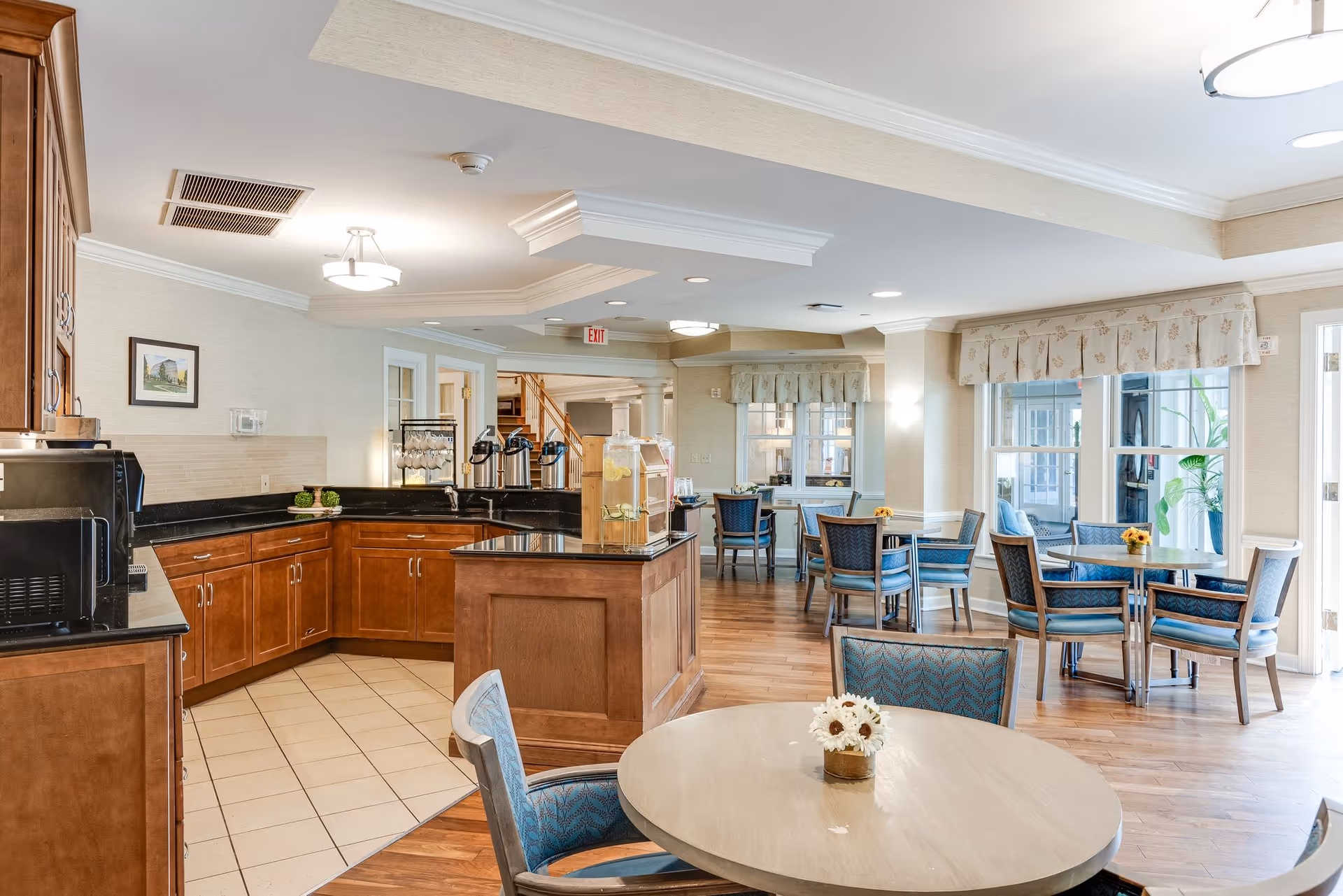 A bright and clean dining area in a senior living facility featuring round tables with blue cushioned chairs, a small kitchen area with wooden cabinets and black countertops, beverage dispensers, and large windows with valances allowing natural light to fill the room.