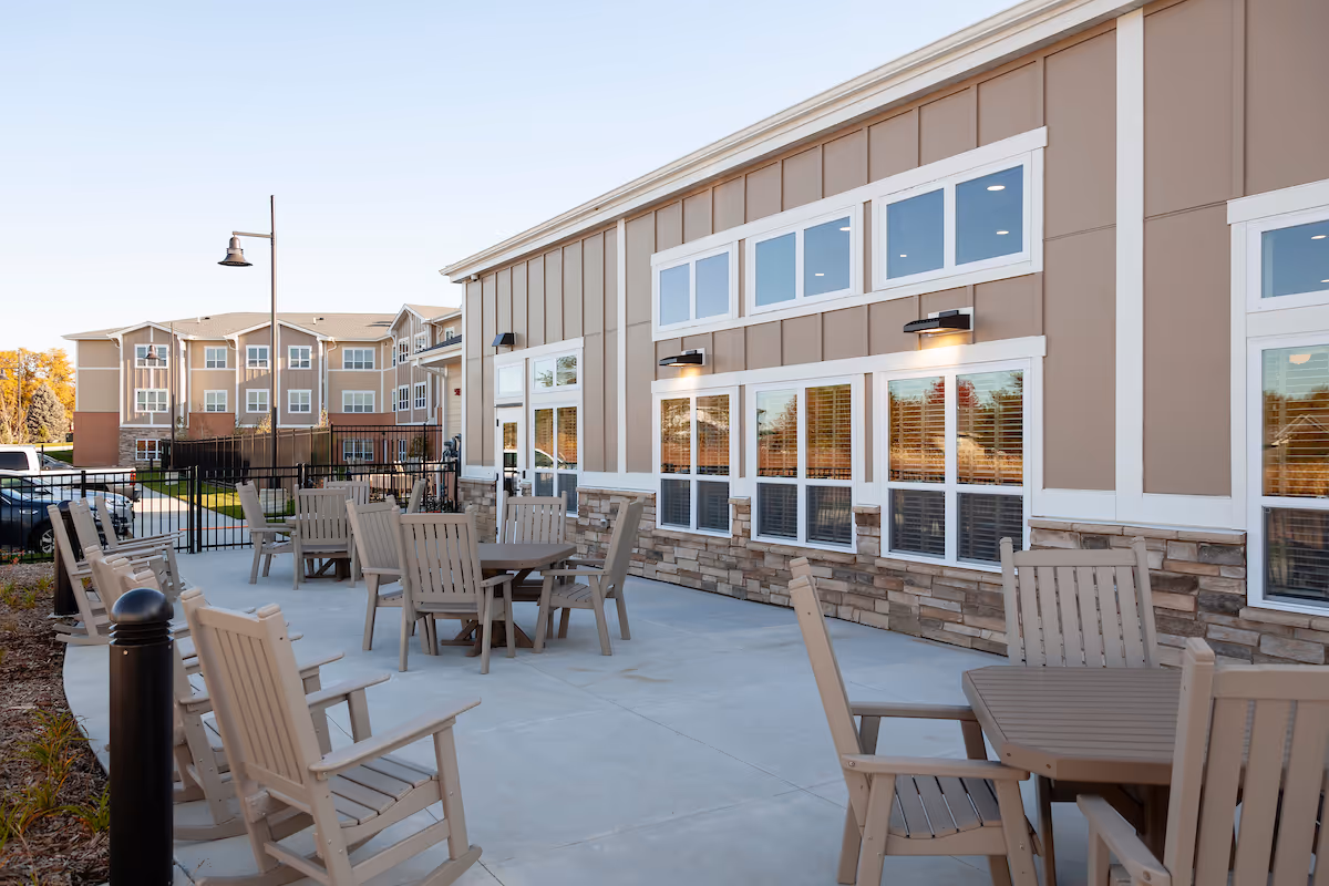 Outdoor patio area at Cedarhurst Senior Living of La Vista with multiple beige wooden tables and chairs arranged on a concrete surface next to a building with large windows and stone accents. The sky is clear and a street lamp is visible in the background.