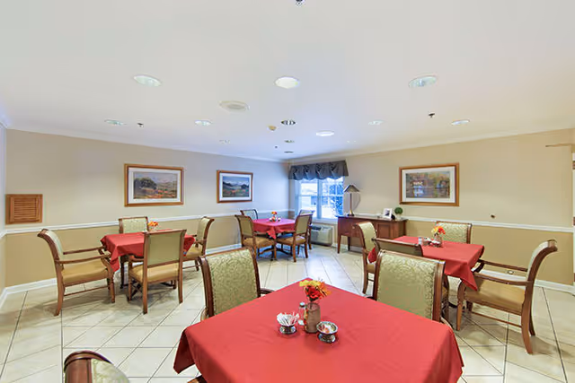 A dining room with several tables covered in red tablecloths, each surrounded by chairs. The room has beige walls with white trim, framed landscape paintings, a window with a valance, and a sideboard with a lamp and decorative items. The floor is tiled in a light color.